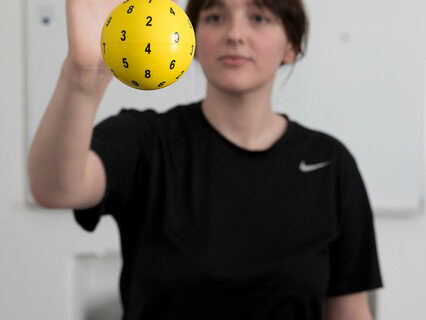 Woman performing a vision therapy exercise by reaching toward a yellow numbered ball suspended in front of her, with a whiteboard in the background.