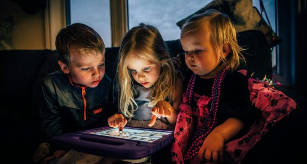 Three young children sitting on a couch, hunched over a bright tablet screen, illustrating screen time and its effect on eyesight
